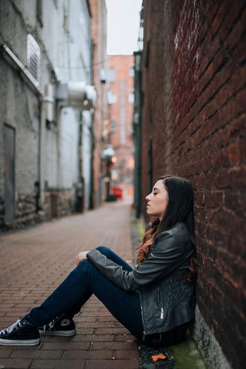 Lexi sitting against a brick wall in an alley, wearing a leather jacket